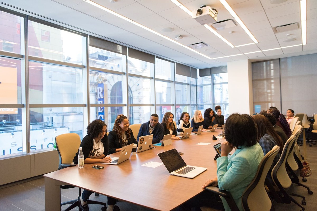 Diverse professionals with laptops around a long conference table beside floor-to-ceiling city windows