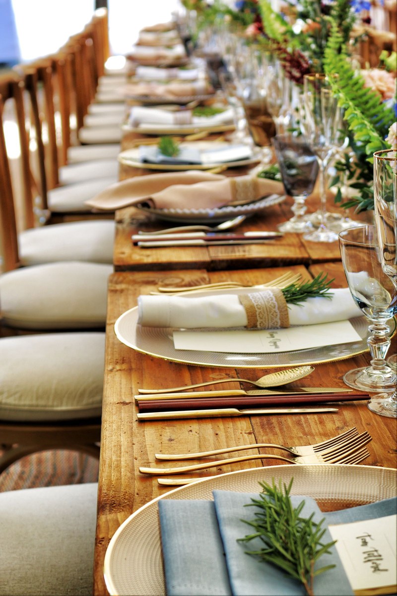 Long rustic wooden banquet table with gold-rimmed plates, gold cutlery, and floral centrepieces